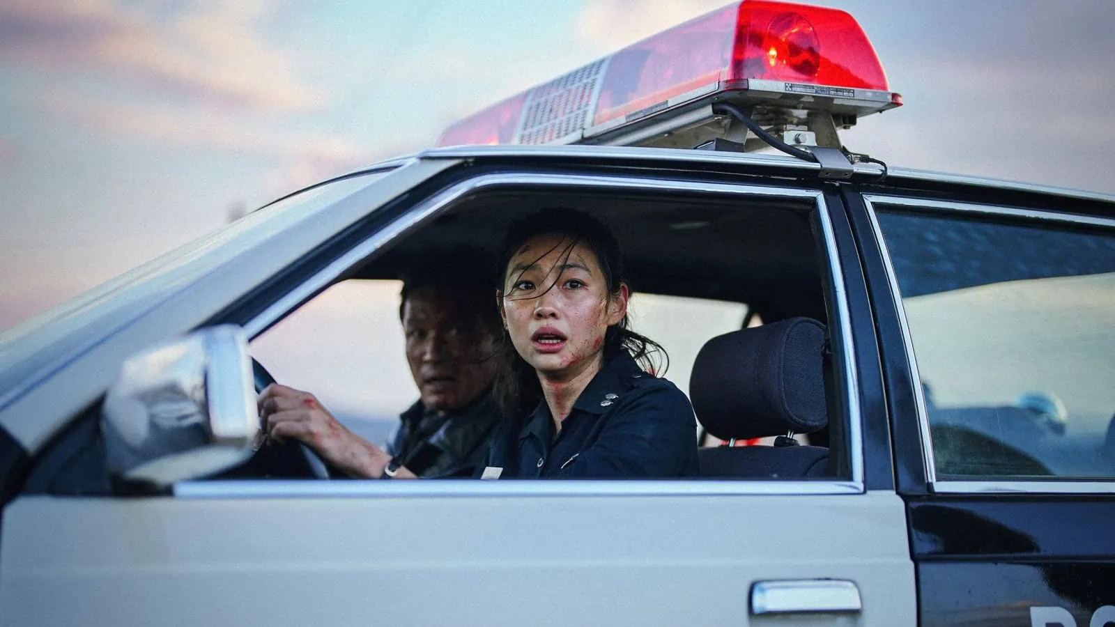 Hoyeon driving a cop car with co-star Jungmin Hwang in the passenger seat.