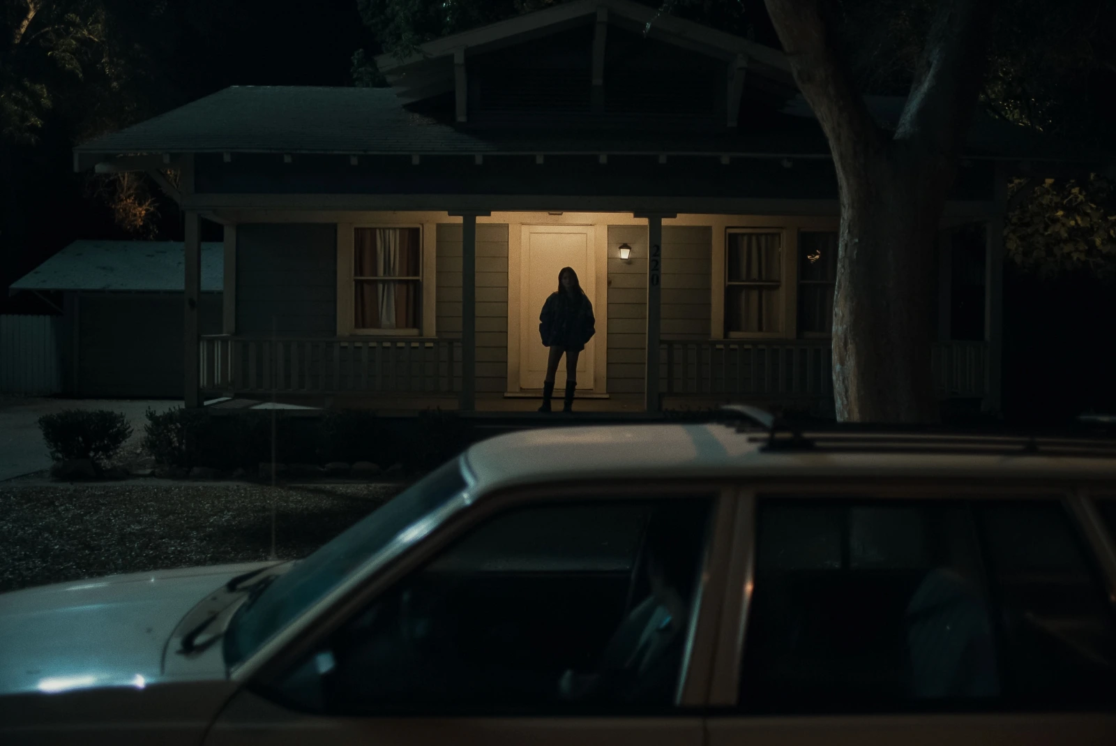 Silhouette of Inde Navarette standing on the porch of a house at night, looking at a car with Michael Johnston in it.