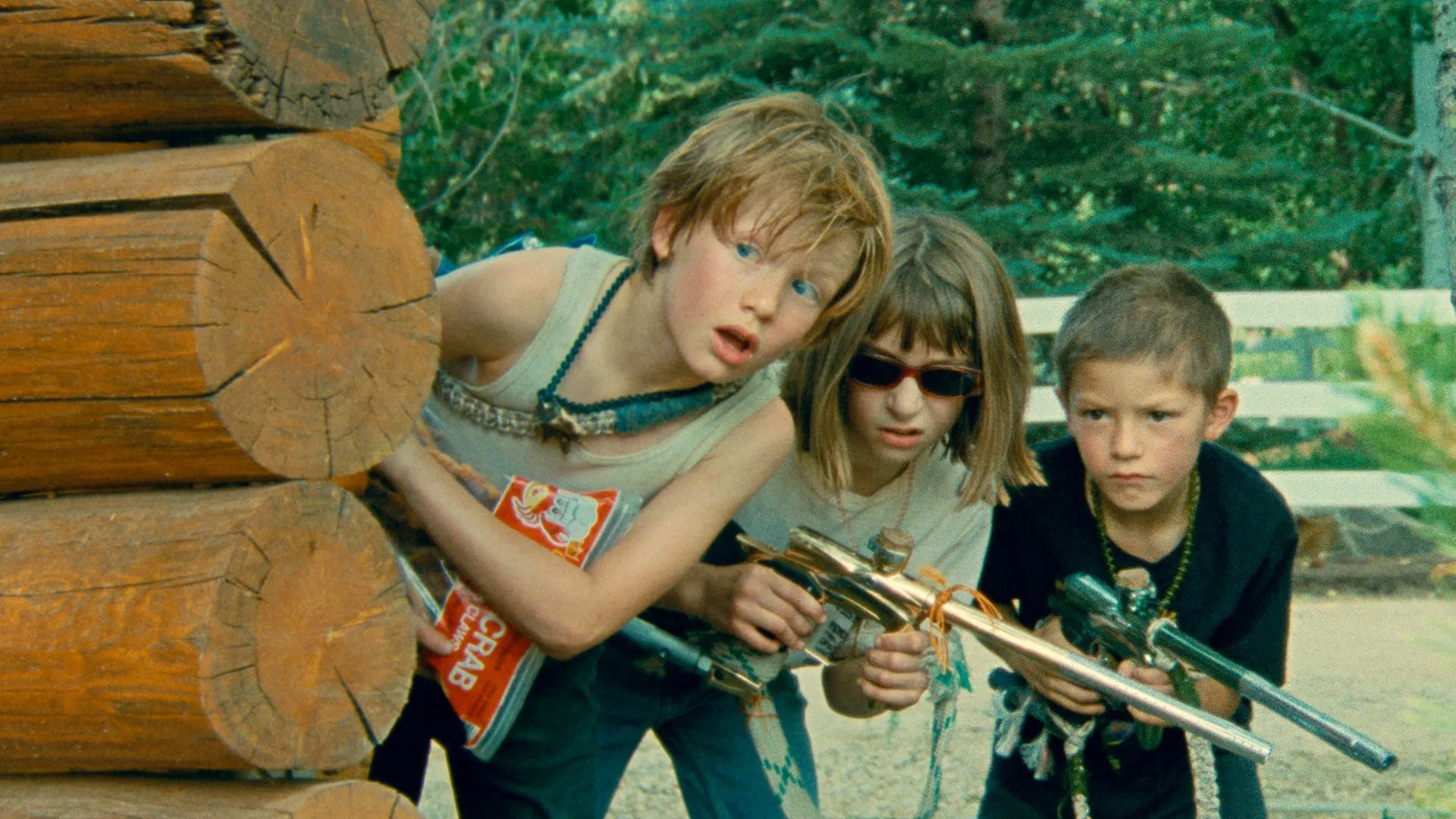 Three young actors looking from behind a pile of wood, holding paint ball guns.