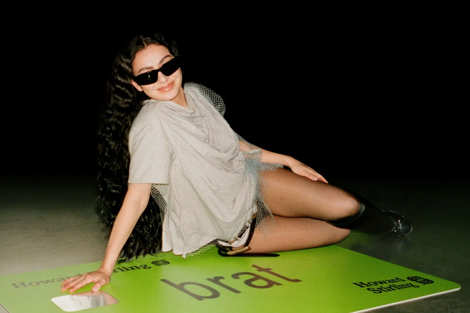Charli XCX sitting on top of a comically large green credit card that says 'brat'.
